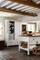 An angled view of a beautiful white linen kitchen featuring a kitchen run with an aged brass Ionian tap, freestanding table, a pantry cupboard, and a collection of vintage furniture and items reflecting on the well-reserved historical space with its original plastered walls, wooden beams and oak floors.