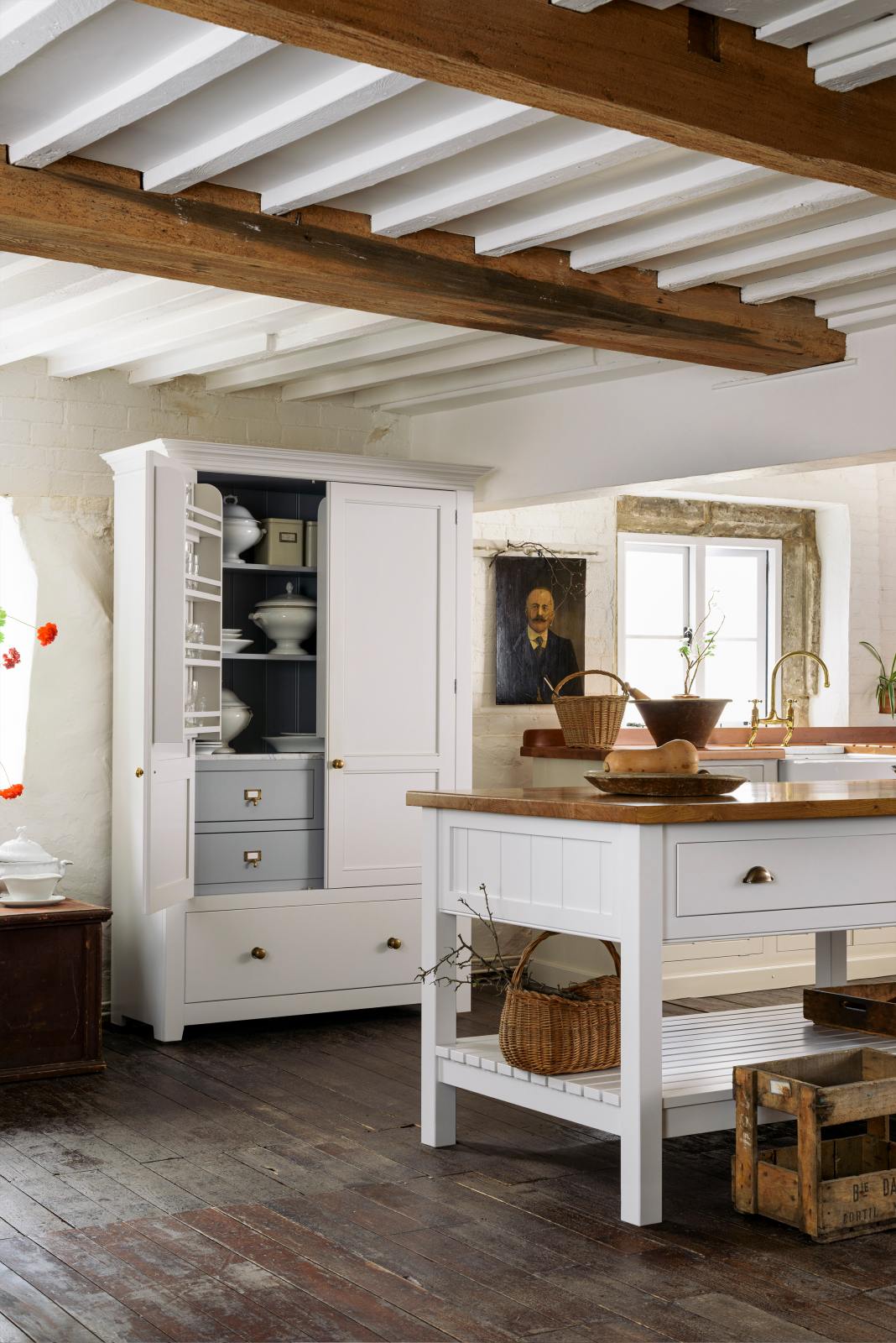 An angled view of a beautiful white linen kitchen featuring a kitchen run with an aged brass Ionian tap, freestanding table, a pantry cupboard, and a collection of vintage furniture and items reflecting on the well-reserved historical space with its original plastered walls, wooden beams and oak floors.