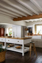 An angled view of a beautiful white linen kitchen featuring a sink run with an aged brass Ionian tap, a freestanding table, a pantry cupboard, and a collection of vintage furniture and items reflecting on the well-reserved historical space with its original plastered walls, wooden beams and oak floors.