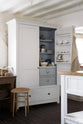 A beautiful linen white kitchen features an open pantry cupboard with a spice rack, marble counter, shelves and closed drawers, and a collection of vintage furniture and items reflecting on the well-reserved historical space with its original plastered walls, wooden beams and oak floors.