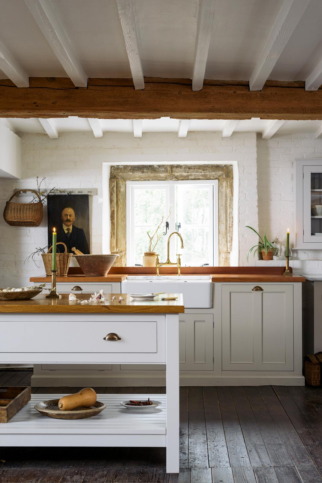 An angled view of a beautiful white linen kitchen featuring a sink run with aged brass Ionian taps, a freestanding table, and a collection of vintage furniture and items reflecting on the well-reserved historical space with its original plastered walls, wooden beams and oak floors.