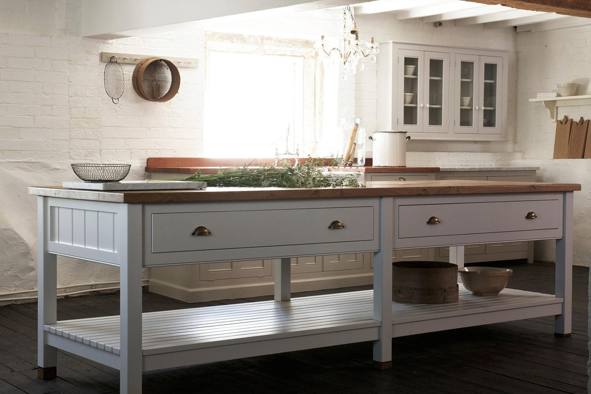 An angled view of a beautiful white linen kitchen featuring a sink run with aged brass Ionian taps, a freestanding table, and a collection of vintage furniture and items reflecting on the well-reserved historical space with its original plastered walls, wooden beams and oak floors.