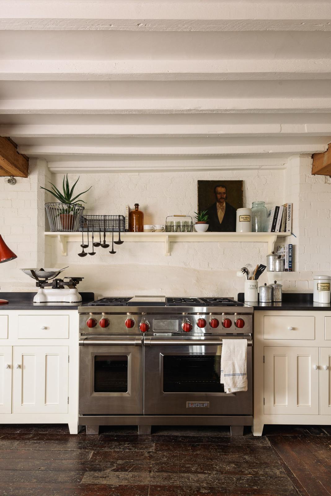 A beautiful white linen kitchen features a cooker run, a silver gas stove, a shaker shelf, and a collection of vintage furniture and items reflecting on the well-reserved historical space with its original plastered walls, wooden beams and oak floors.