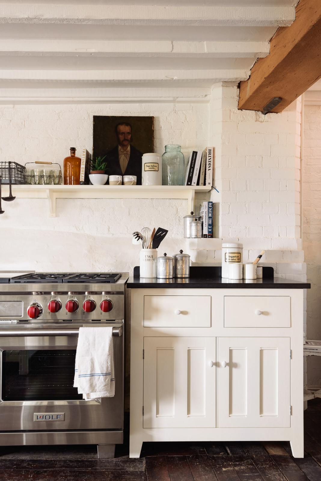 A beautiful white linen kitchen features a cooker run, a silver gas stove, a shaker shelf, and a collection of vintage furniture and items reflecting on the well-reserved historical space with its original plastered walls, wooden beams and oak floors.
