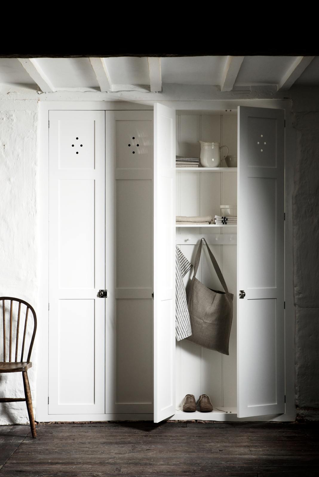 A beautiful linen white kitchen features a built-in pantry cupboard with high shelves, a peg rail, and a collection of vintage furniture and items reflecting on the well-reserved historical space with its original plastered walls, wooden beams and oak floors.
