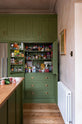 Nestled between the oak worktop kitchen island and the entrance of a romantic, decorative, Parisian-inspired classic English green kitchen is a large, open full-length cabinet with closed top cupboards and a built-in stainless steel fridge-freezer. The cabinet is fully stocked with sauces and spices in spice racks, shelves of dried ingredients, and a mixer. It's adorned with a bouquet of blush-brown chrysanthemums, lace runners, and a wooden-framed artwork.
