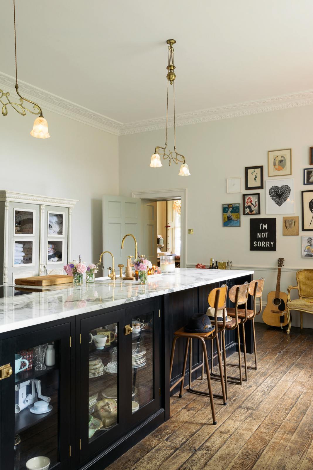 An oblique view of a tall-ceilinged classic English kitchen centres around a wide painter’s black island with an aged brass Ionian tap, marble worktop, glazed base cabinets, and a cosy breakfast nook with vintage stools. The space feels artfully lived-in—funky paintings, a guitar, and kitchen essentials tell the story of a creative, full-of-life family.