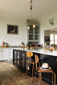 An oblique view of a tall-ceilinged classic English kitchen centres around a wide painter’s black island with an aged brass Ionian tap, marble worktop, glazed base cabinets, and a cosy breakfast nook with vintage stools. The space feels artfully lived-in—funky paintings, hanging sculptures, and kitchen essentials tell the story of a creative, full-of-life family.