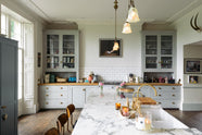 A tall-ceilinged, classic English kitchen features a wide painter’s black island with an aged brass Ionian tap, marble worktop, a cosy breakfast nook with vintage stools, an off-white cooker run, and matching wall cabinets. The space feels artfully lived-in — funky paintings, hanging sculptures, delicate glass gaselier light, and kitchen essentials tell the story of a creative, lively family.