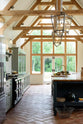 This oak-framed barn kitchen features a long cooker run with a black granite worktop, a bank of glazed larders, a shelf, and a kitchen island complete with a single Belfast sink and an aged brass Ionian tap atop a solid oak worktop. Adorned with framed pendant lights, artisan ceramics, books, and pumpkins, this kitchen creates a warm and inviting space.