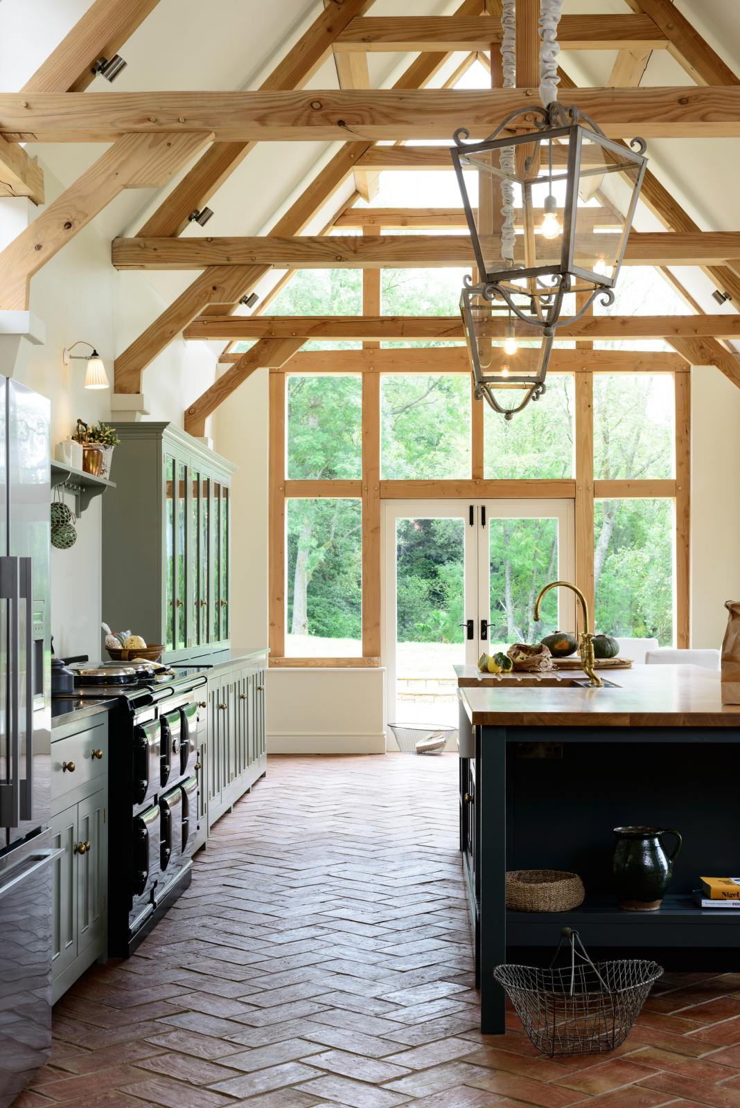 This oak-framed barn kitchen features a long cooker run with a black granite worktop, a bank of glazed larders, a shelf, and a kitchen island complete with a single Belfast sink and an aged brass Ionian tap atop a solid oak worktop. Adorned with framed pendant lights, artisan ceramics, books, and pumpkins, this kitchen creates a warm and inviting space.