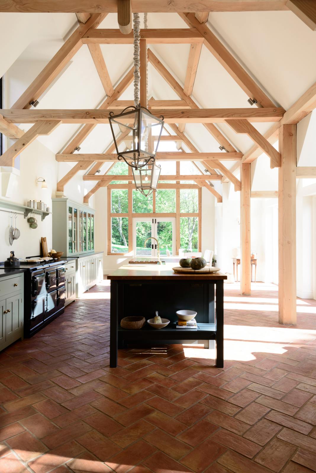 An oblique view of an oak-framed barn kitchen features a long cooker run with a black granite worktop, a bank of glazed larders, a shelf, and a kitchen island complete with a single Belfast sink and an aged brass Ionian tap atop a solid oak worktop. Adorned with framed pendant lights, artisan ceramics, books, and pumpkins, this kitchen creates a warm and inviting space.