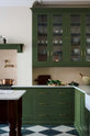 A spacious and elegant Wilkes green classic English kitchen features a long L-shaped cooker and sink run with a Silestone worktop, glazed wall cupboards, shelves, and an Heirloom dairy table. Adorned with classic marble chequered-board flooring, Heirloom task lights, cream subway splashback tiles, copper pots, and luscious green vegetables, it creates a classic and stylish kitchen.