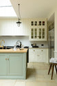 A quiet corner of a bright and spacious classic English kitchen features a long white cooker run with glazed wall cupboards and a pale-blue kitchen island that includes a single Belfast sink, an aged brass Ionian tap, and a solid oak worktop. Adorned with metal embellishments like industrial pendant lights, a silver refrigerator, a jade-green vase filled with a floral arrangement, and vintage wooden furniture, it creates a wonderful mix of simplicity and charismatic personality.
