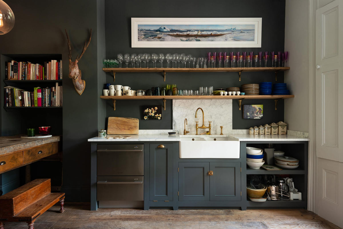 An eclectic, navy blue, classic English kitchen features a short and humble sink run with a double Belfast sink, an aged brass Ionian tap, a Carrara marble worktop, and a square splashback at the centre. The long, corridor-like space is beautifully decorated with beloved vintage items and furniture, paintings, a mixed-media deer wall hanging and unique kitchen equipment, giving the room a lot of whimsy, character and functionality.