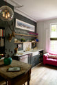 An angled view of an eclectic, navy blue, classic English kitchen features a short and humble sink run with a double Belfast sink, an aged brass Ionian tap, a Carrara marble worktop, and a square splashback at the centre. The long, corridor-like space is beautifully decorated with beloved vintage items and furniture, paintings, a mixed-media deer wall hanging, and a vibrant couch, giving the room a lot of whimsy, character and functionality.