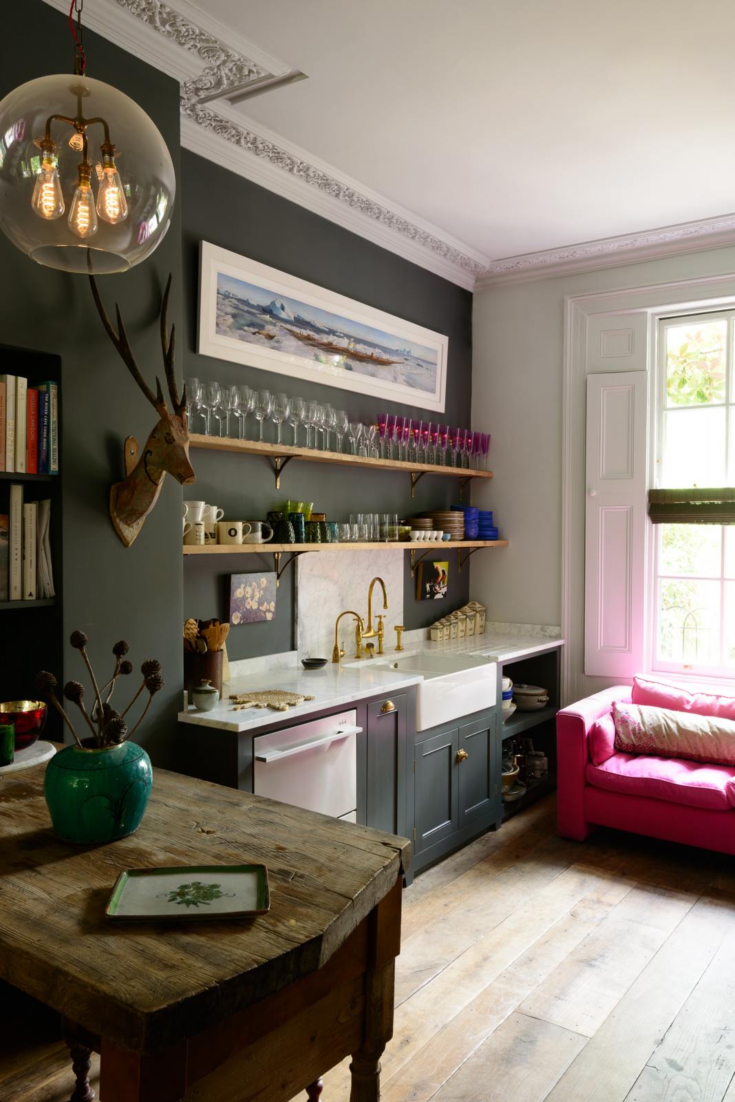 An angled view of an eclectic, navy blue, classic English kitchen features a short and humble sink run with a double Belfast sink, an aged brass Ionian tap, a Carrara marble worktop, and a square splashback at the centre. The long, corridor-like space is beautifully decorated with beloved vintage items and furniture, paintings, a mixed-media deer wall hanging, and a vibrant couch, giving the room a lot of whimsy, character and functionality.