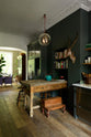 An eclectic, navy blue, classic English kitchen features a freestanding cabinet with a large silver fridge and glazed cabinets. The long, corridor-like space is beautifully decorated with beloved vintage items and furniture, paintings, a mixed-media deer wall hanging and unique lighting, giving the room a lot of whimsy, character and functionality.