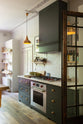 An eclectic, navy blue, classic English kitchen features a short and humble cooker run with a silver gas cooker, shaker canopy with a Carrara marble worktop, and a square splashback at the centre. The long, corridor-like space is beautifully decorated with beloved vintage items and furniture, paintings, copper pendant lights and unique kitchen equipment, giving the room a lot of whimsy, character and functionality.