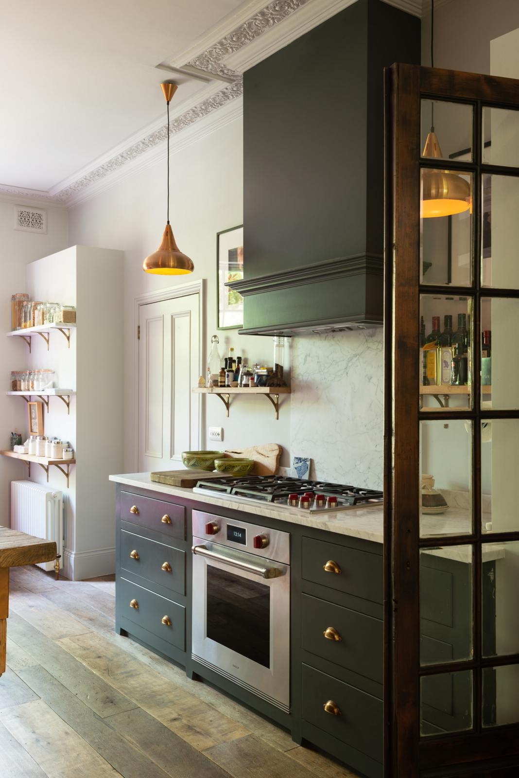 An eclectic, navy blue, classic English kitchen features a short and humble cooker run with a silver gas cooker, shaker canopy with a Carrara marble worktop, and a square splashback at the centre. The long, corridor-like space is beautifully decorated with beloved vintage items and furniture, paintings, copper pendant lights and unique kitchen equipment, giving the room a lot of whimsy, character and functionality.