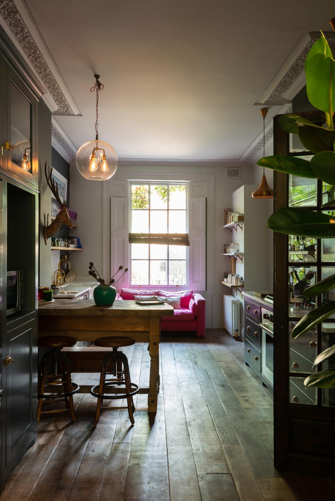 An eclectic, navy blue, classic English kitchen features a short and humble sink run, cooker run and a freestanding cabinet. The long, corridor-like space of a Georgian townhouse is beautifully decorated with beloved vintage items and furniture, paintings, copper pendant lights and unique kitchen equipment, giving the room a lot of whimsy, character and functionality.