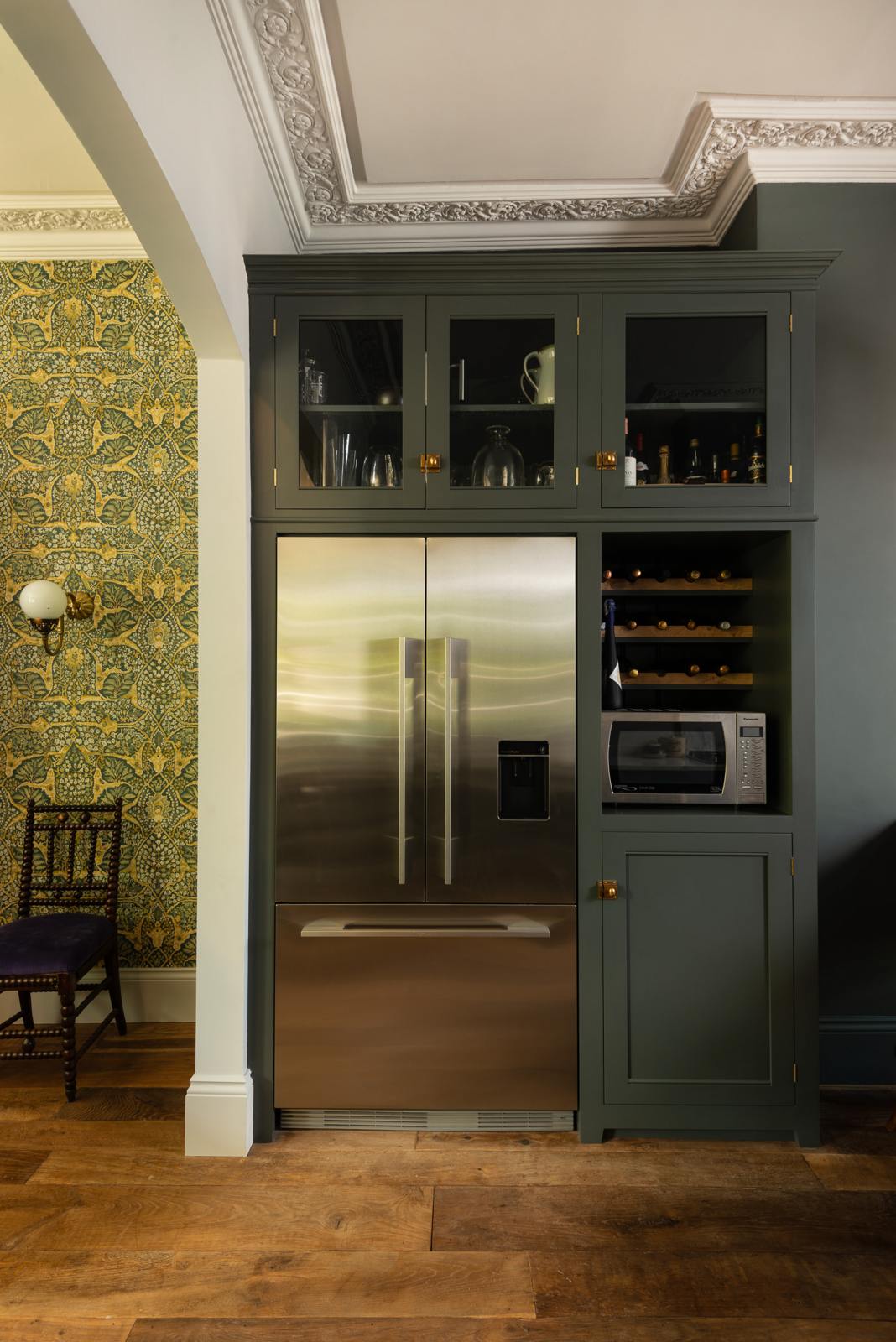 An eclectic, navy blue, classic English kitchen features a freestanding cabinet with a large silver fridge and glazed cabinets. The long, corridor-like space is beautifully decorated with beloved vintage items and furniture, a wine bottle rack, and a maximalist wallpaper in the dining room, giving the room a lot of whimsy, character and functionality.