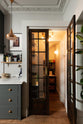 An eclectic, navy blue, classic English kitchen features a small pantry closet using a set of vintage doors with their original brass pull handles. The long, corridor-like space is beautifully decorated with beloved vintage items and furniture, and a copper pendant light, giving the room a lot of whimsy, character and functionality.