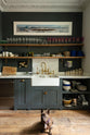 An eclectic, navy blue, classic English kitchen features a short and humble sink run with a double Belfast sink, an aged brass Ionian tap, a Carrara marble worktop, and a square splashback at the centre. The long, corridor-like space is beautifully decorated with beloved vintage items and furniture, paintings, unique kitchen equipment and a cute small dog, giving the room a lot of whimsy, character and functionality.