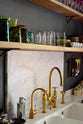 A closer look at the eclectic, navy blue, classic English kitchen’s sink run, with a double Belfast sink, an aged brass Ionian tap, a Carrara marble worktop, and a square splashback at the centre. The long, corridor-like space is beautifully decorated with beloved vintage items and furniture, paintings, and unique kitchen equipment, giving the room a lot of whimsy, character and functionality.