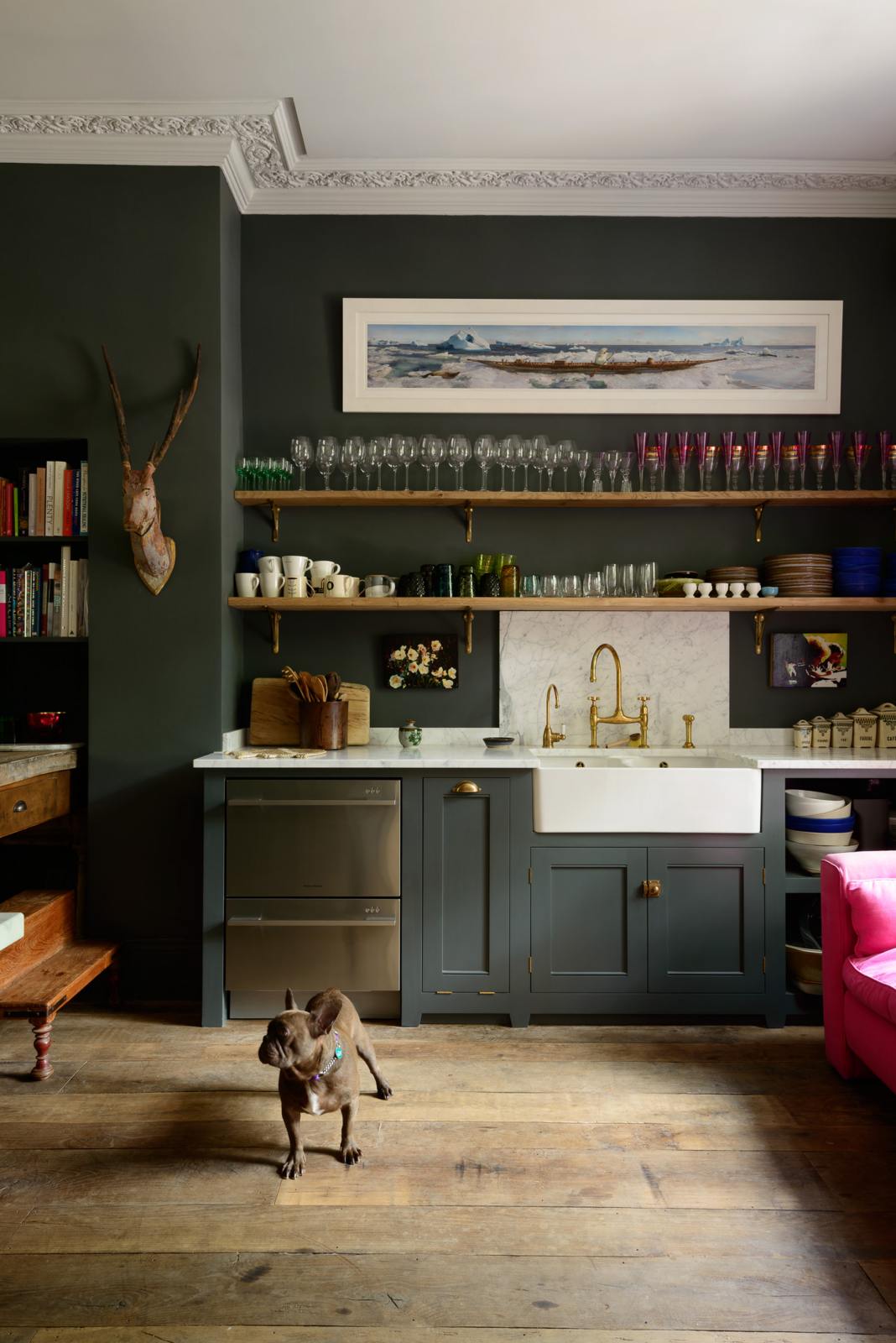 An eclectic, navy blue, classic English kitchen features a short and humble sink run with a double Belfast sink, an aged brass Ionian tap, a Carrara marble worktop, and a square splashback at the centre. The long, corridor-like space is beautifully decorated with beloved vintage items and furniture, a mixed-media deer wall hanging, unique kitchen equipment and a cute dog, giving the room a lot of whimsy, character and functionality.