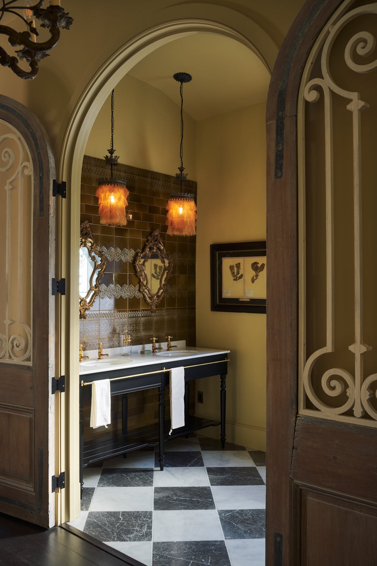 Looking past the antique Parisian doors into a beautiful butter-yellow powder room, you’ll find a black twin Victorian washstand, tall sepia lace market tiles, and a Parisian chequered marble floor. Paired with antique mirrors, pendant lights, and framed pressed botanical artworks, the room exudes a glamorous, vintage charm.
