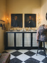An oblique view of a beautiful butter-yellow powder room, you’ll find a pair of Habedasher’s glazed base cabinets with ruched curtains, a collection of antique furniture, vintage paintings, and a Parisian chequered marble floor. Paired with floral wall candle holders and an old dress form, the room exudes a glamorous, vintage charm.