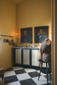 An oblique view of a beautiful butter-yellow powder room, you’ll find a pair of Habedasher’s glazed base cabinets with ruched curtains, a collection of antique furniture, vintage paintings, and a Parisian chequered marble floor. Paired with floral wall candle holders and an old dress form, the room exudes a glamorous, vintage charm.