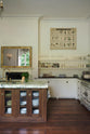 A grand yet simple white linen classic English kitchen, featuring white and cream splashback tiles, a cooker run with a polished sapele worktop, and a kitchen island topped with Arabescato marble, a polished brass sink, and an aged brass Ionian tap. The space is decorated with a large scientific-style illustration of different vegetable species and a large metal-and-crystal chandelier.