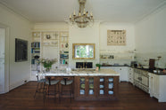 A grand yet simple linen-white classic English kitchen, featuring white and cream splashback tiles, a cooker run with a polished sapele worktop, and a kitchen island topped with Arabescato marble, a polished brass sink, and an aged brass Ionian tap. The space is simply decorated with a large scientific-style illustration of different vegetable species, a glass vase filled with wildflowers from the area, and a large metal-and-crystal chandelier.