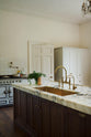 A closer look at the kitchen island reveals a surface topped with Arabescato marble, a polished brass sink, and an aged brass Ionian tap—all part of a grand yet simple linen-white classic English kitchen. The space is decorated with a garnish of parsley beside a butternut squash, and a large metal-and-crystal chandelier.