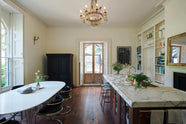 In a grand yet simple linen-white classic English kitchen, one sees an Arabescato marble kitchen island, a polished brass sink, an aged brass Ionian tap, a large dining table with metal-framed chairs upholstered in black cushions, and a vintage French door leading to the garden. The space is simply decorated with books, glass vases filled with wildflowers from the area, and a large metal-and-crystal chandelier.