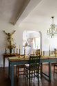 A gorgeous dining room features a teal Millhouse refectory table with vintage wooden chairs, furniture, a large variety of brass candlesticks, a crystal chandelier and a Jesus statue on the wall. And through the open archway is a simple and functional linen white scullery room with a modest sink run with polished sapele, a large single Carrara marble sink, and a deVOL laundry maid.
