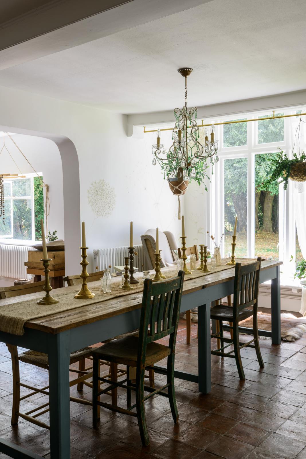 A beautiful dining room features a teal Millhouse refectory table with vintage wooden chairs, an array of brass candlesticks, a crystal chandelier, and a stunning view of the mill pond and the English countryside.