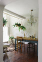 Looking through an open archway is a beautiful dining room with a teal Millhouse refectory table, vintage wooden chairs, an array of brass candlesticks, a crystal chandelier, hanging plants and an intricate wild flower illustration wallpaper.