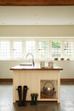 This simple and spacious classic English kitchen features a kitchen island equipped with a double Belfast sink and an antique silver Ionian tap atop a solid oak worktop. Adorned with a vintage metal basket, simple ceramics, copper pans, and boots, it creates a humble and functional family kitchen.