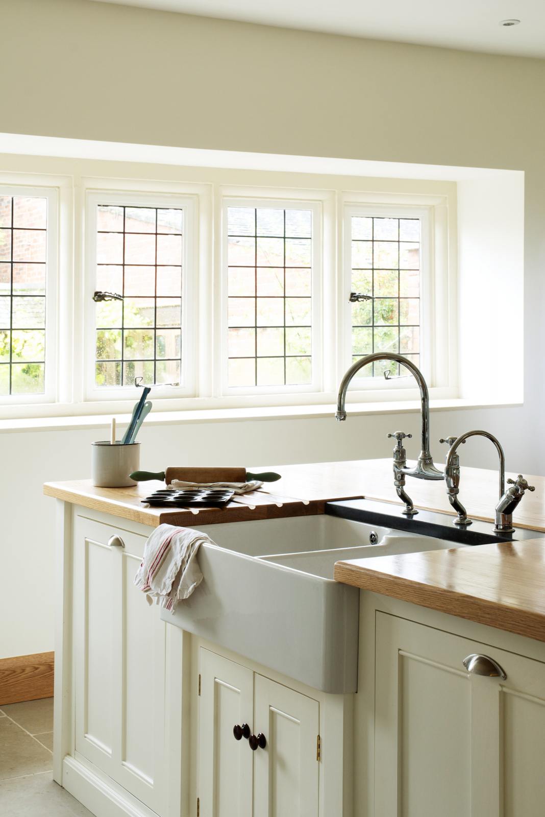 An oblique view of a simple and spacious classic English kitchen features a kitchen island with a double Belfast sink and an antique silver Ionian tap atop a solid oak worktop. Adorned with simple ceramics, a backing tray, tea towels, and a rolling pin, it creates a humble and functional family kitchen.