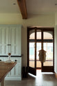 This simple and spacious classic English kitchen features a kitchen island with a solid oak worktop, a double Belfast sink, an antique silver Ionian tap, and a free-standing closed larder pantry cupboard. Adorned with a glass decanter featuring a metal funnel and a vest hanging on the door handle, this kitchen creates a humble and stylish space.