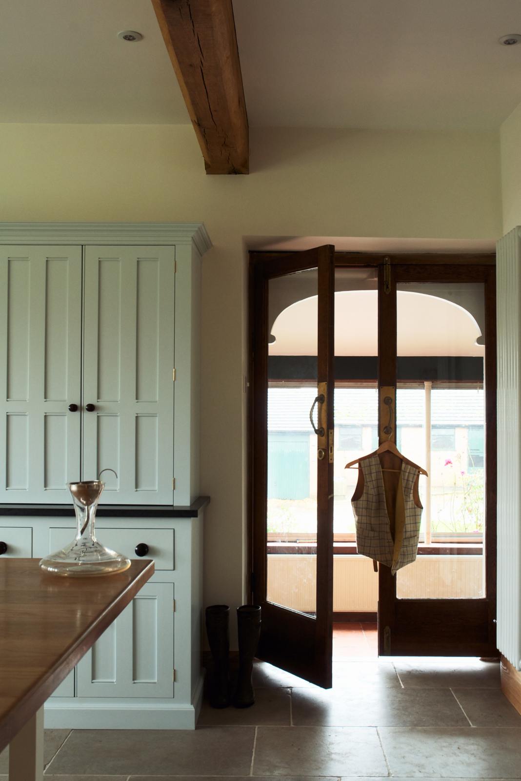 This simple and spacious classic English kitchen features a kitchen island with a solid oak worktop, a double Belfast sink, an antique silver Ionian tap, and a free-standing closed larder pantry cupboard. Adorned with a glass decanter featuring a metal funnel and a vest hanging on the door handle, this kitchen creates a humble and stylish space.