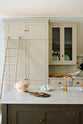 One side of a grand, earth-toned classic kitchen features a Silestone worktop cooker run, glazed wall cupboards, a full-height pantry, a large island with a copper worktop, and a ladder. Adorned with warm colours such as an aged brass hanging rail, wooden chartulary board, vintage ceramics, dried pasta and eggs, it makes a spectacular use of space but is also down-to-earth.