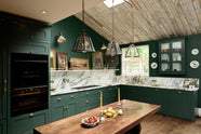 A lush green, classic English kitchen featuring an L-shaped sink and cooker, all complemented by Arabescato marble for the worktop, splashback, and countertop shelf. The kitchen includes full-height cabinets and a wall cupboard, creating a luxurious yet bohemian atmosphere. An angled wooden skylight window allows natural light to flow into the room, highlighting delicate bird-painted plates, a rich oak dining table, framed antique paintings, and industrial-style gas light fixtures on the walls.