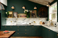 A lush green, classic English kitchen featuring an L-shaped sink and cooker, all complemented by Arabescato marble for the worktop, splashback, and countertop shelf. An angled wooden skylight window allows natural light to flow into the room, highlighting white lilies in glass vases, a rich oak dining table, framed antique paintings, and industrial-style gas light fixtures on the walls.