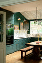 A lush green, classic English kitchen featuring an L-shaped sink and cooker, all complemented by Arabescato marble for the worktop, splashback, and countertop shelf. The kitchen includes full-height cabinets and a wall cupboard, creating a luxurious yet bohemian atmosphere. An angled wooden skylight window allows natural light to flow into the room, highlighting a rich oak dining table with a matching bench, framed antique paintings, and industrial-style gas light fixtures on the walls.