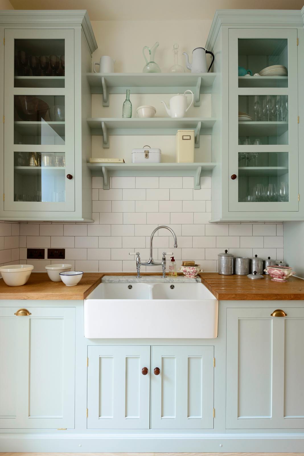 A close-up of a pale-blue classic English galley kitchen features a sink run with a solid oak worktop, a double Belfast sink, a chrome Ionian tap, glazed wall cupboards, pantry cupboards, and shelves. Adorned with artisan ceramics, vintage containers, and simple platters, it creates a humble and practical kitchen that embraces vintage 80s aesthetics.