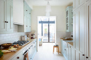This pale-blue classic English galley kitchen features a cooker and sink run with a solid oak worktop, a double Belfast sink, a chrome Ionian tap, wall cupboards, a canopy, and shelves. Adorned with a vintage scale, a bread tin, artisan ceramics, silver containers, and colourful vegetables, it creates a humble and practical kitchen that embraces vintage 80s aesthetics.